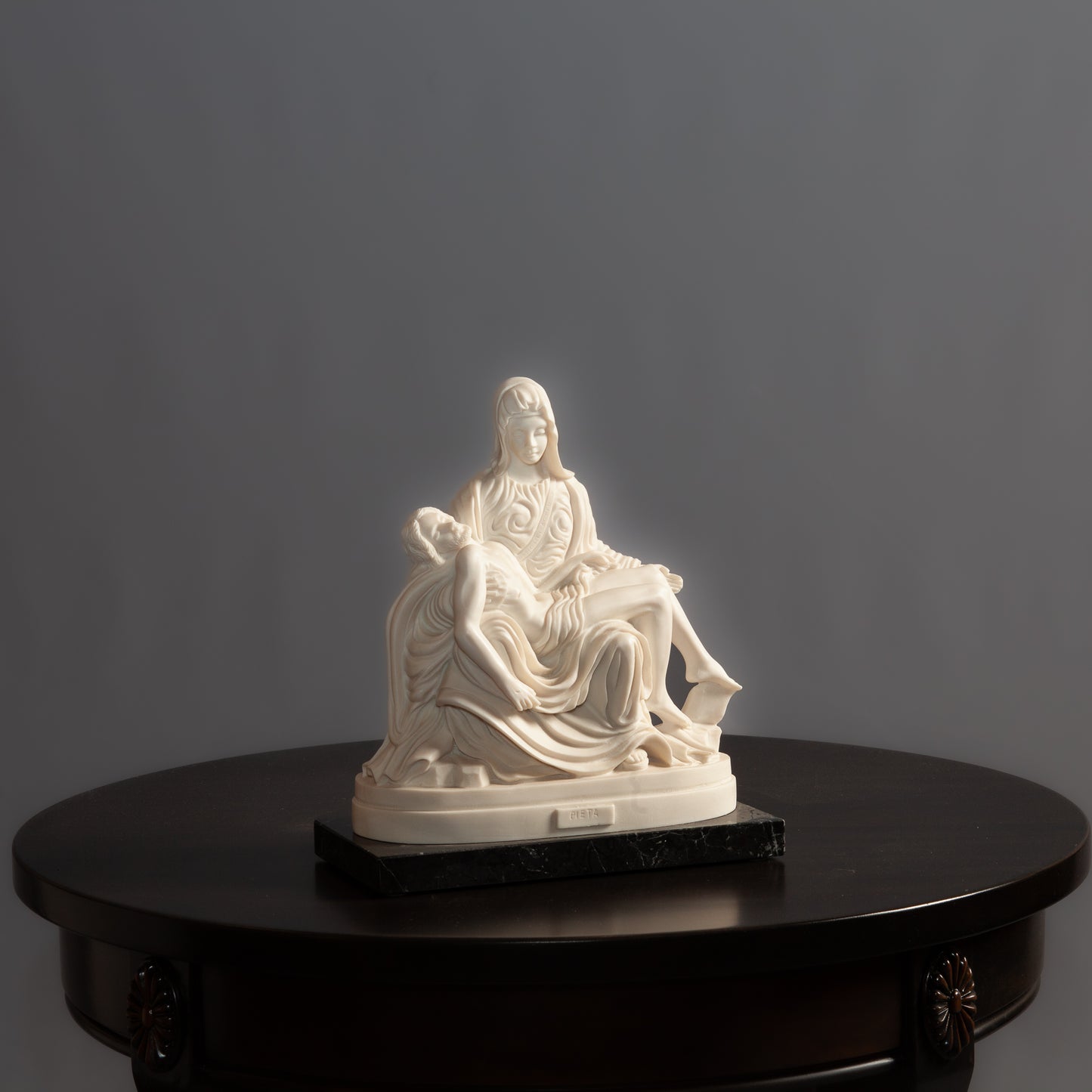 A white Pietà sculpture on a black marble base sits on a dark wooden table against a gray background.