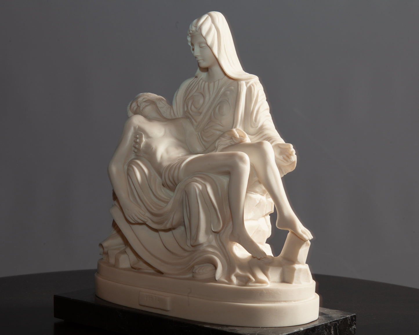 Side view of white Pietà sculpture depicting Mary holding Jesus rests on a black marble base atop a dark wooden table.