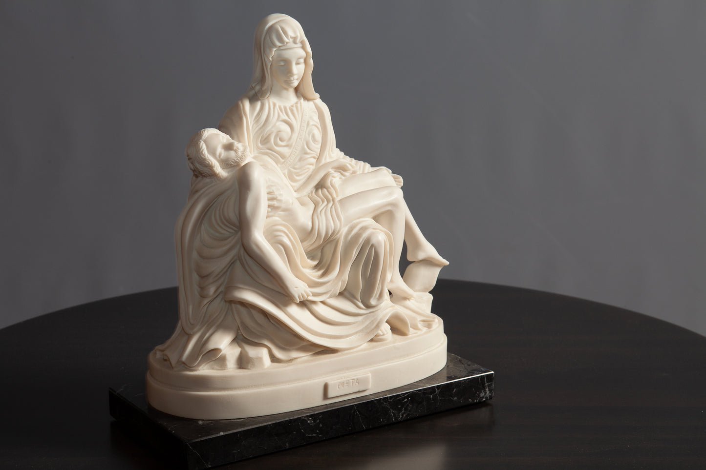 A white Pietà sculpture depicting Mary holding Jesus rests on a black marble base atop a dark wooden table.