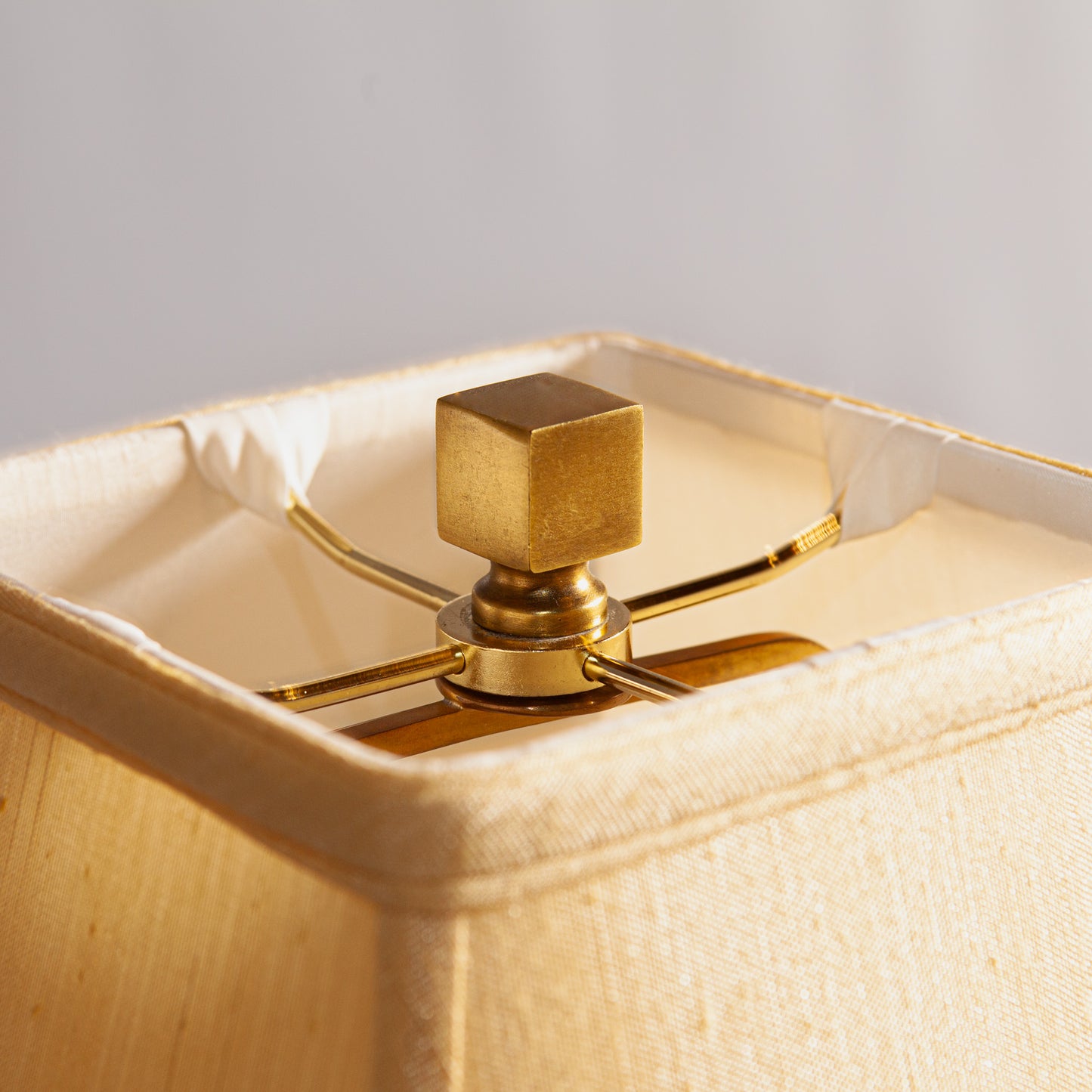 A close-up of a beige lampshade with a gold square finial and metal frame.