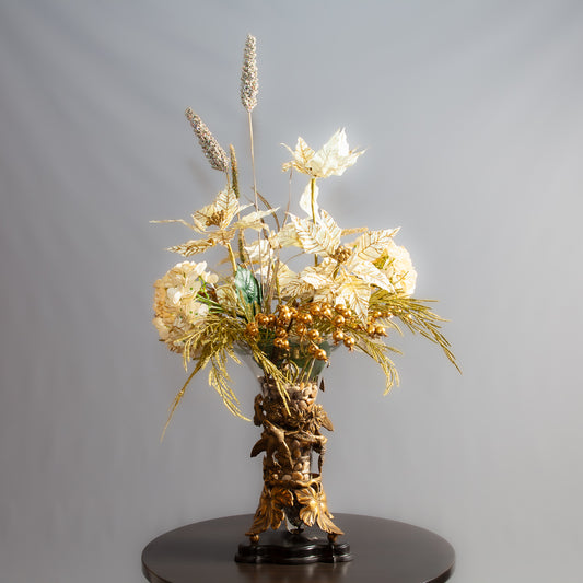 A decorative gold floral arrangement with intricate metallic leaves and flowers sits in an ornate vase on a table.