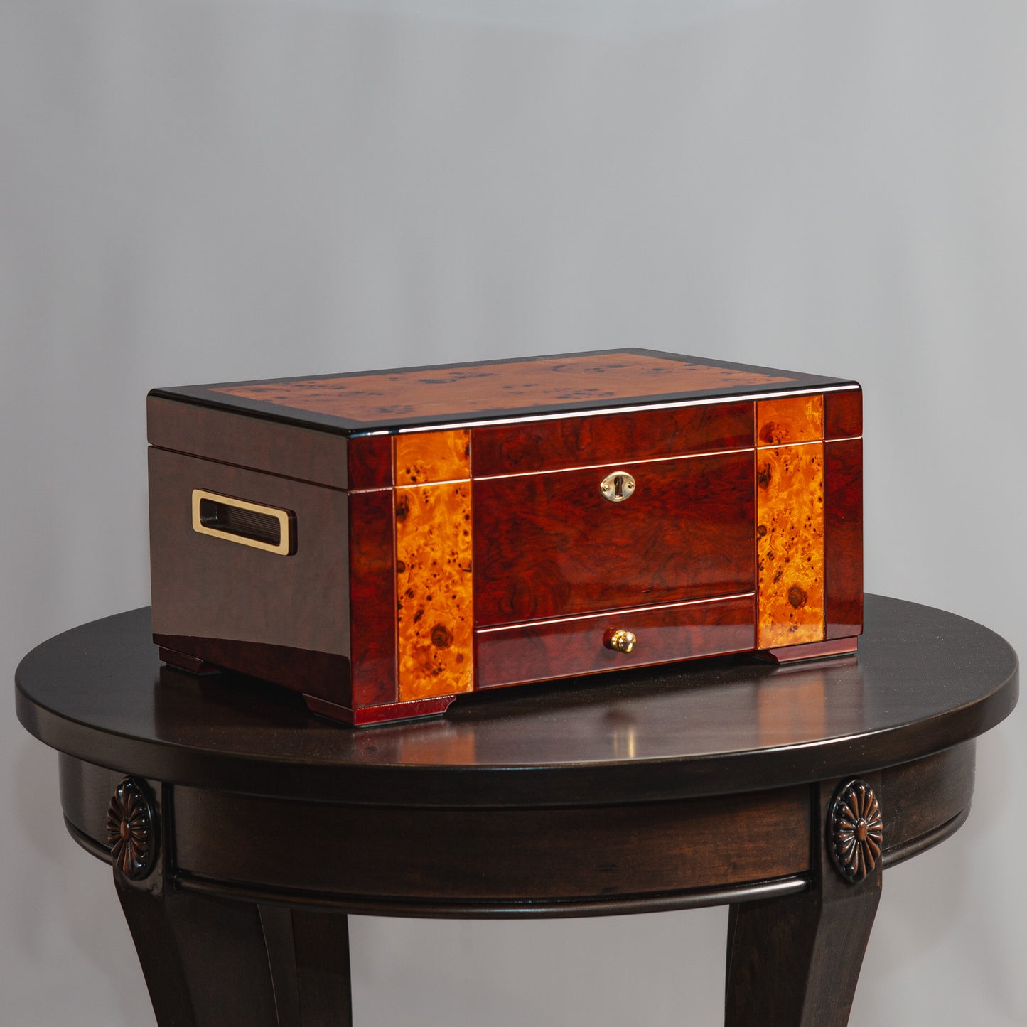 A polished wooden jewelry box with gold accents and burl wood inlays sits on a dark wooden table.