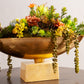 A gold decorative bowl with vibrant succulents and greenery sits on a marble pedestal base