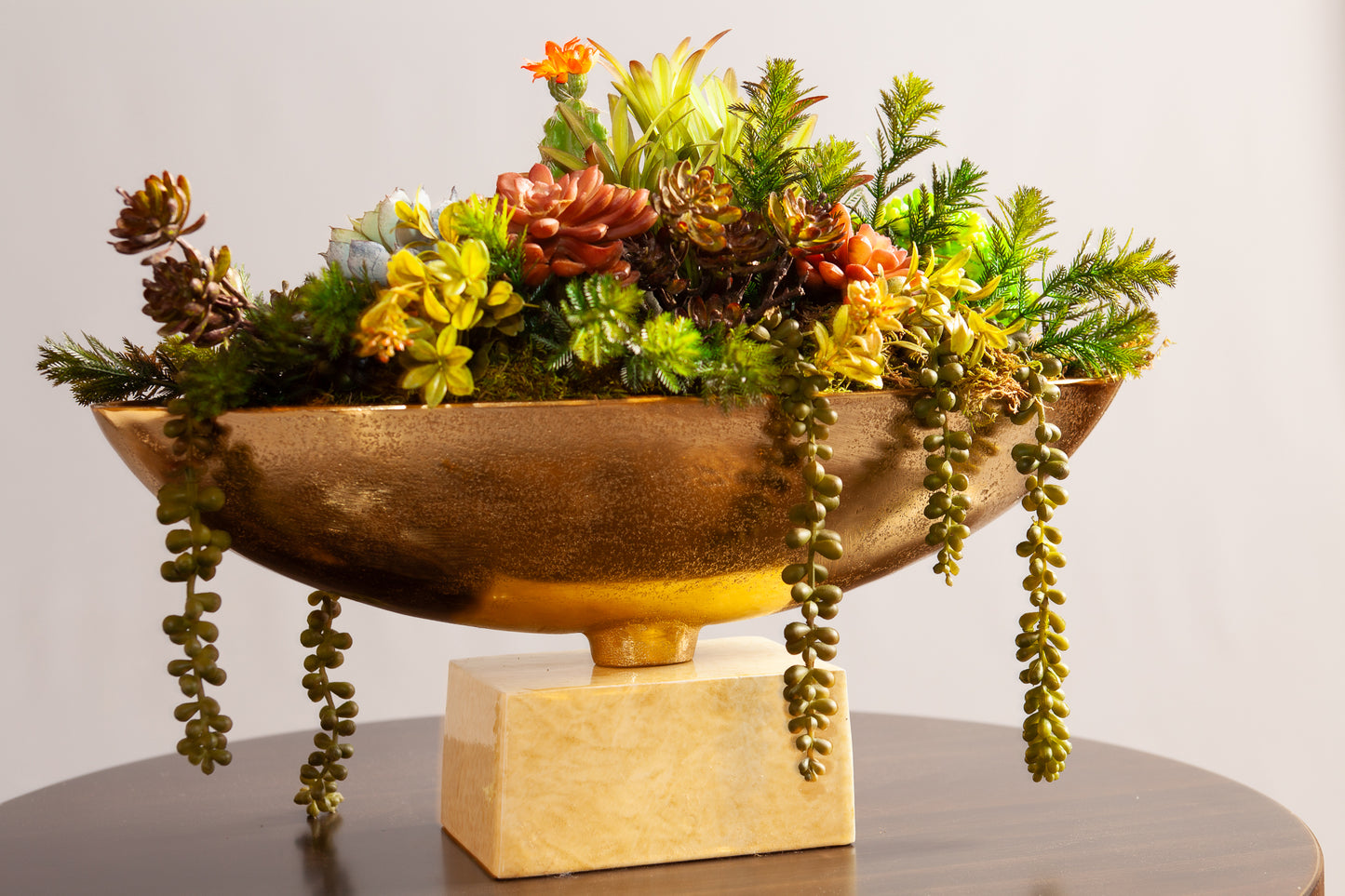 A gold decorative bowl with vibrant succulents and greenery sits on a marble pedestal base